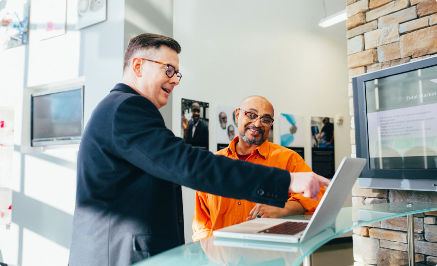 Two men in suits standing beside a laptop, engaged in a discussion or collaboration about chatbots